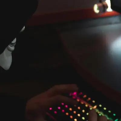 Stereotype Hacker with black hoodie, guy fawkes mask sitting infront of a simulated computer screen with his hands typing on a rgb backlit keyboard