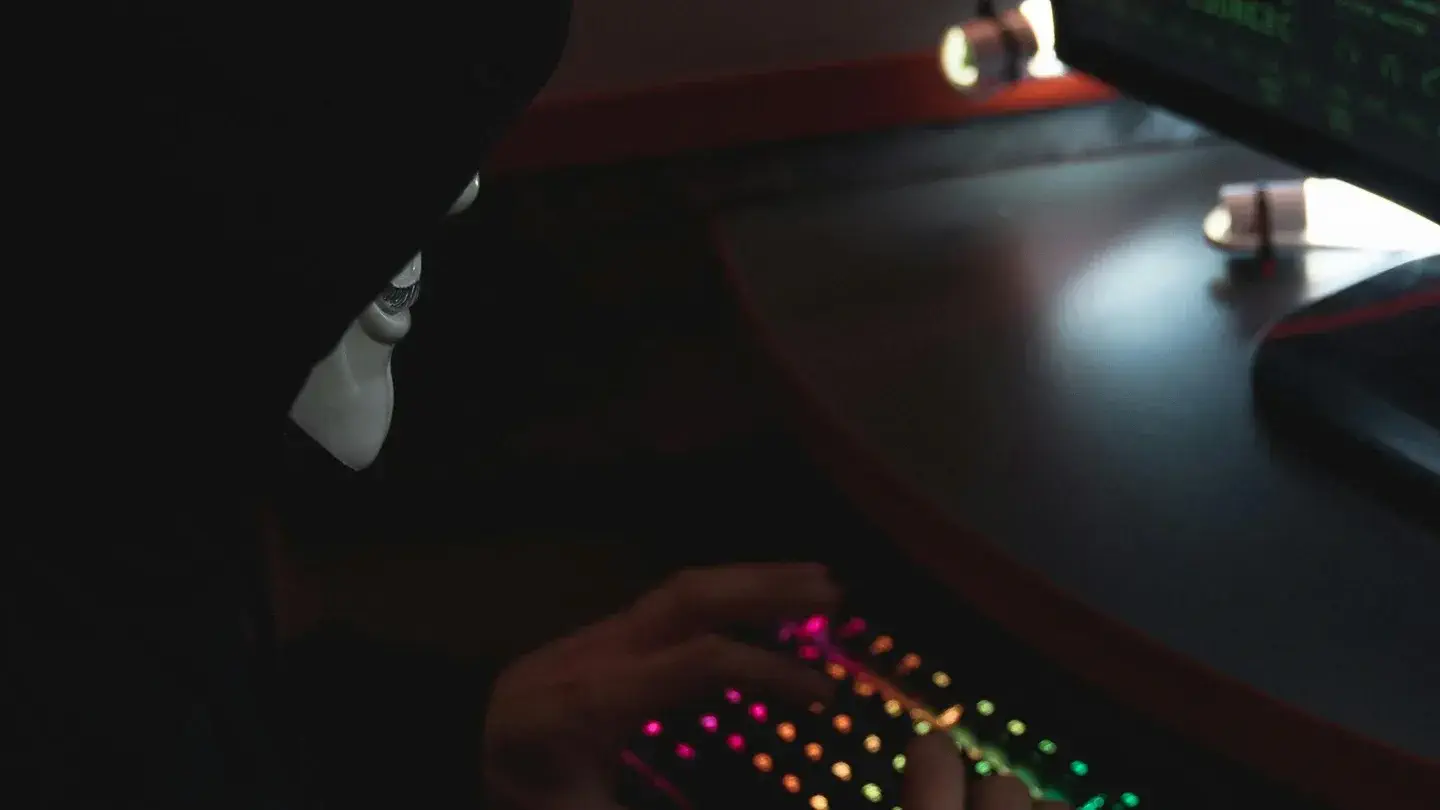 Stereotype Hacker with black hoodie, guy fawkes mask sitting infront of a simulated computer screen with his hands typing on a rgb backlit keyboard